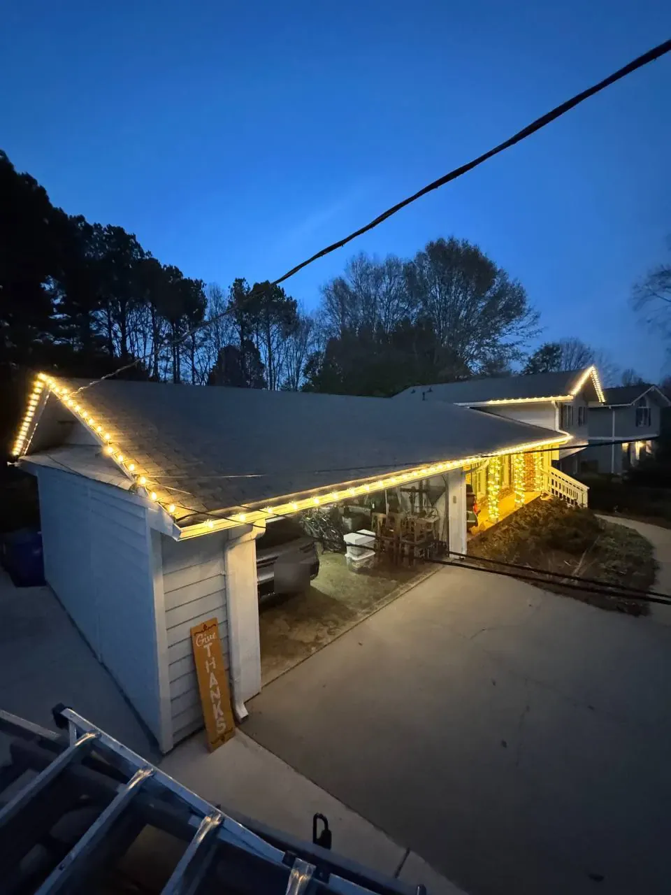 Warm-white lights along a ranch roofline, seen from the top of the installer's ladder at dusk